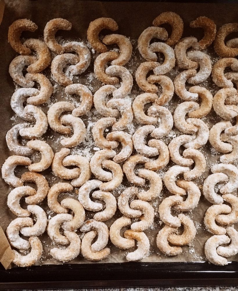 Ein Blech mit goldbraunen Vanillekipferln. Aneinander gereiht, dass sie fast so aussehen wie eine Kette. Sie liegen auf braunem Backpapier. Zwischen den Kipferln liegt verteilt etwas Puderzucker.