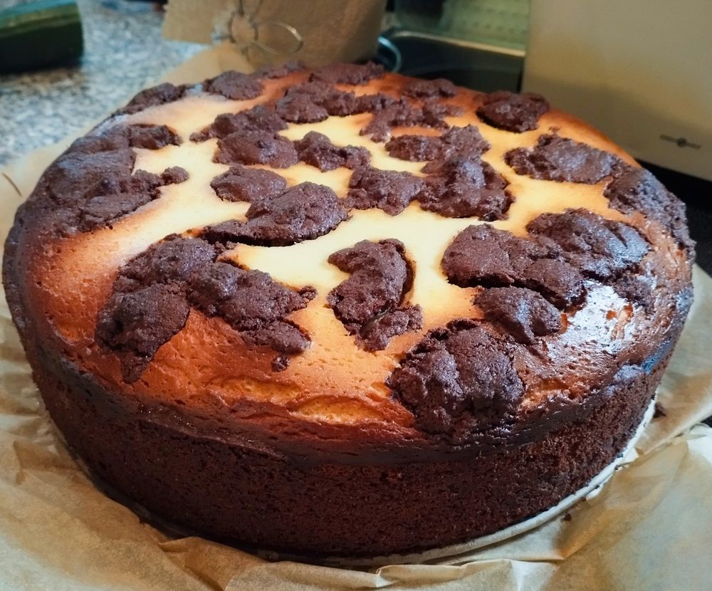 Auf hellbraunem Backpapier steht ein großer runder Zupfkuchen. Oben verteilt auf dem Kuchen wahrlos gezupfte dunkelbraune Teigfetzen. Umso näher man in die Mitte des Kuchens schaut, umso heller wird der Käsekuchenteig. Im Hintergrund erkennt man unscharf einen Teil einer grünen Salatgurke und den unteren Teil einer Küchenpapierrolle. Ebenso erkennt man auch noch ein Teil des Ceranfeldes, auf dem der Kuchen steht, nebst der Arbeitsplatte. 
Persönliche Meinung: 
Der Kuchen sieht wirklich lecker und einladend aus. Was man nicht sieht ist, dass im inneren noch Pflaumen verarbeitet sind, die dem Kuchen eine fruchtige Note verleihen. Alles versteckt unter einer goldbraunen Schicht aus Käsekuchen mit Flecken und Fetzen des Teigs vom Boden und Rand. 
