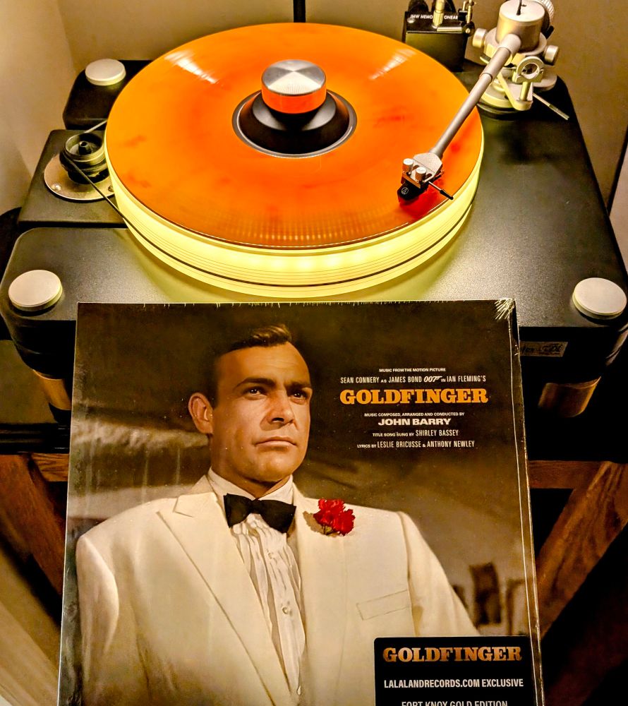 John Barry – Goldfinger (Music From The Motion Picture) on colored vinyl (yellow orange splatter) Playing on VPI Scoutmaster turntable, 12-pound acrylic platter glowing yellow. Audio Technica AT-VM95ML cartridge.

Album cover is photo of Sean Connery as James Bond in a white tuxedo
