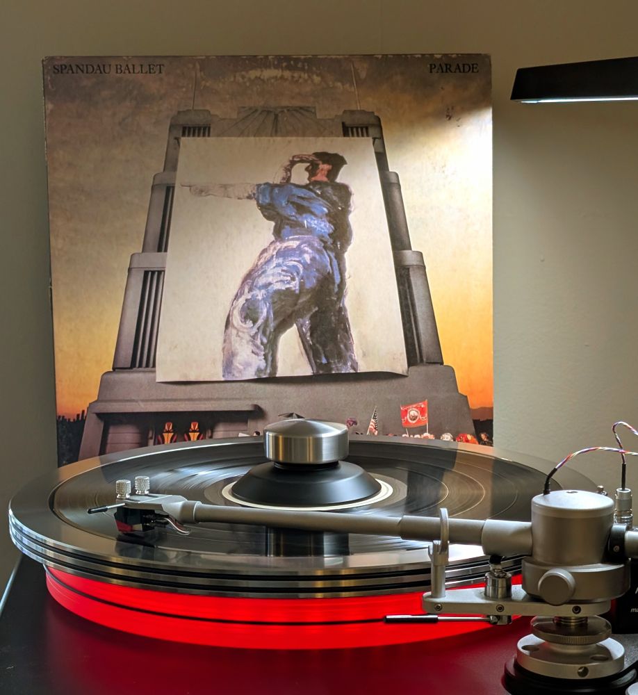 Spandau Ballet 'Parade' vinyl LP playing on VPI Scoutmaster turntable, 12-pound acrylic platter glowing red. Audio Technica AT-VM95ML cartridge.
Album cover (from Wikipedia) is a "painted banner of the profile of a man standing and pointing straight ahead with one hand while the other provides a visor for his eyes. The man represented their experience during the True tour of moving forward while being blinded by a shower of accolades.[16][k] The banner is prominently displayed on the album cover, where it hangs in front of an art deco building.

A diverse procession makes its way below the banner, on the street level, in keeping with the parade theme."
