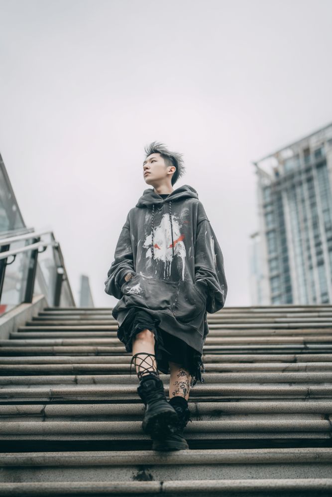 A young person walks down a wide outdoor staircase in an urban setting, framed dramatically from a low angle that emphasizes their stride and posture. The sky above is pale and overcast, casting a diffuse gray light that blends with the muted tones of the cityscape. The subject wears an oversized, distressed dark hoodie splattered with white and red paint-like patterns, the hood draping heavily over their frame. Their hair is styled in sharp, spiked strands that catch the wind, giving a sense of motion. Black boots with intricate lace-up details and visible tattoos on their leg add to the raw, rebellious energy of their look. The concrete steps stretch down toward the viewer, while glass railings and the blurred structure of a high-rise building loom at the edges, suggesting a modern city environment. The overall atmosphere is moody and cinematic, balancing grit with quiet confidence.

Keywords:
urban fashion, streetwear, staircase, oversized hoodie, moody sky, rebellious style, cityscape, tattoos, black boots, dramatic angle, modern architecture, cinematic mood

Created using Midjourney and my Echoform™ prompt structure. The original render was saved as a JPEG in Lightroom to optimize file size. This is a complete, unedited image. Use is permitted for non-commercial creative projects.