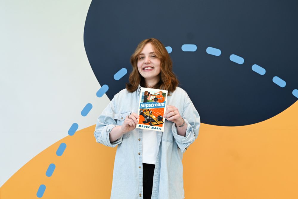 Author Madge Maril holding her book, "Slipstream," in front of an art wall at the Downtown Main Library.