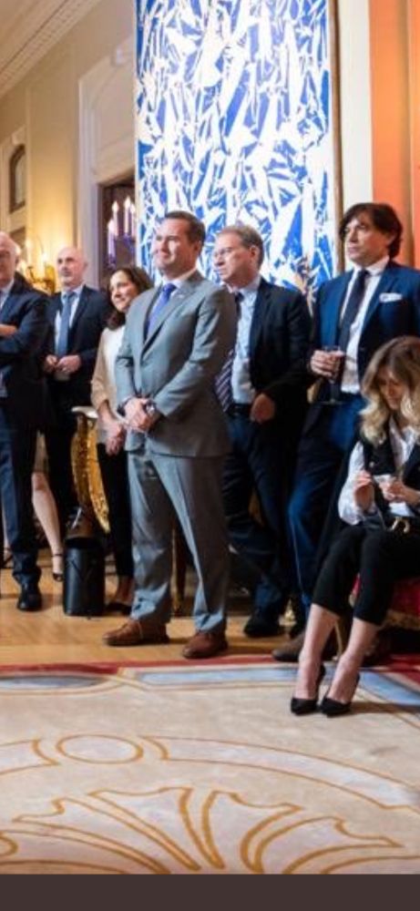 Photo of waltz standing next to Goldberg at event at the French embassy in 2021 Both are wearing suits, standing amongst a crowd of people as they watch a person with a microphone speak