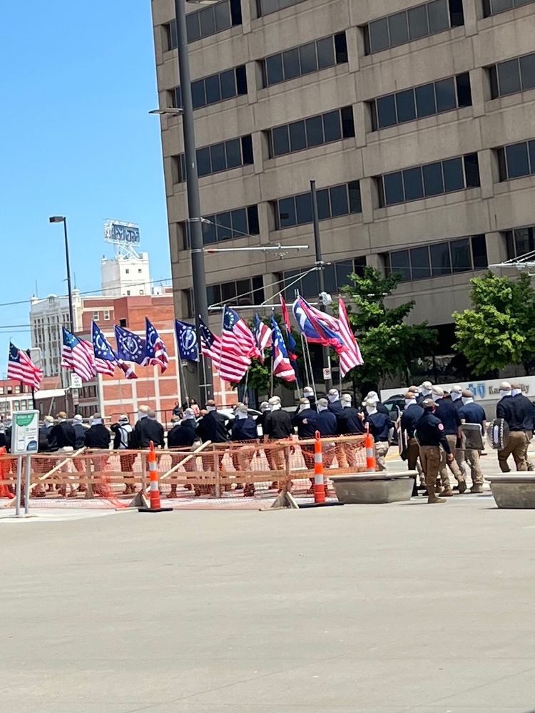 Group of men in khakis, navy shirts, and masks walking in the city center of Kansas City, Mo. they’re carrying flags 