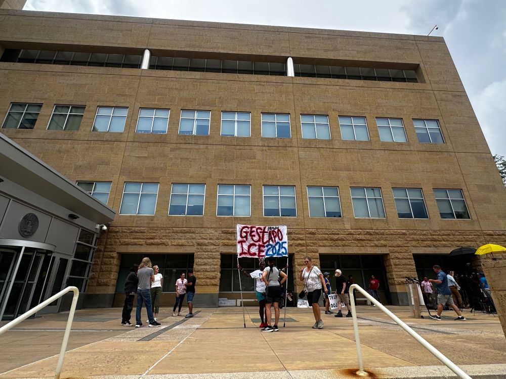Federal courthouse in greenbelt with people milling about outside. A woman holds a sign that reads "Gestapo ICE 2025"