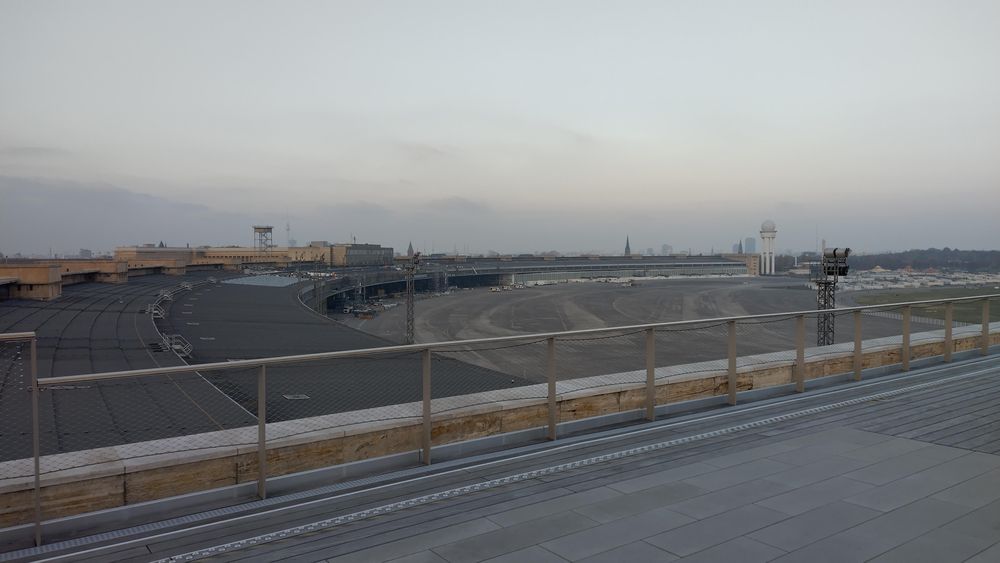 Blick auf das Vorfeld. Eine große Betonfläche vor dem Flughafengebäude und seinen Hangars. Im Hintergrund der Fernsehturm.