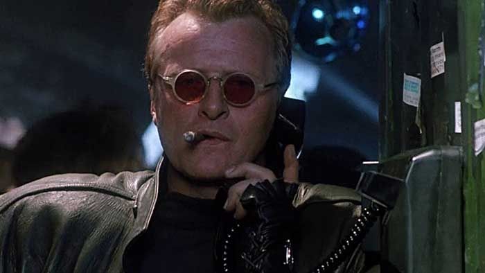 Hauer in Split Second, chomping on a cigar, wearing tiny little red-tinted round sunglasses, fingerless gloves, a future leather jacket, talking into a retro-future payphone, his power-alley hair up medium high.