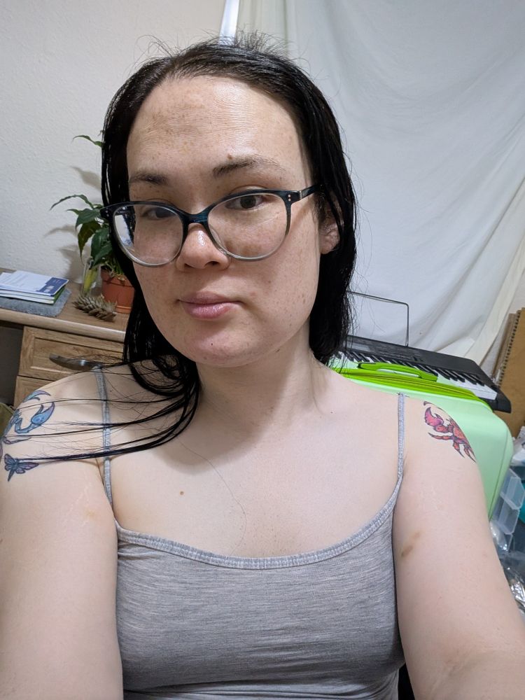 Selfie of a woman wearing a grey cami top with her keyboard and plant in the background, and her shoulder tattoos partially visible