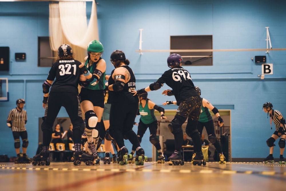 Full wide action shot of a pack. The tall skater wearing a green Nottingham Roller Derby crop top, short black shorts and a green helmet cover with a star being, and full skate gear, is trying to get through a wall of two skaters on black uniforms, with skaters from both teams in action in the background