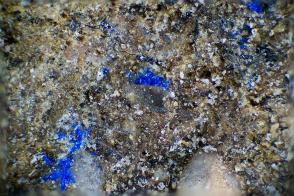 Microscopic photo of the blue spots, that are irregular in shape and size and positioned diagonally across the image. The rest of the photo shows the rough sand coloured texture of the stone.