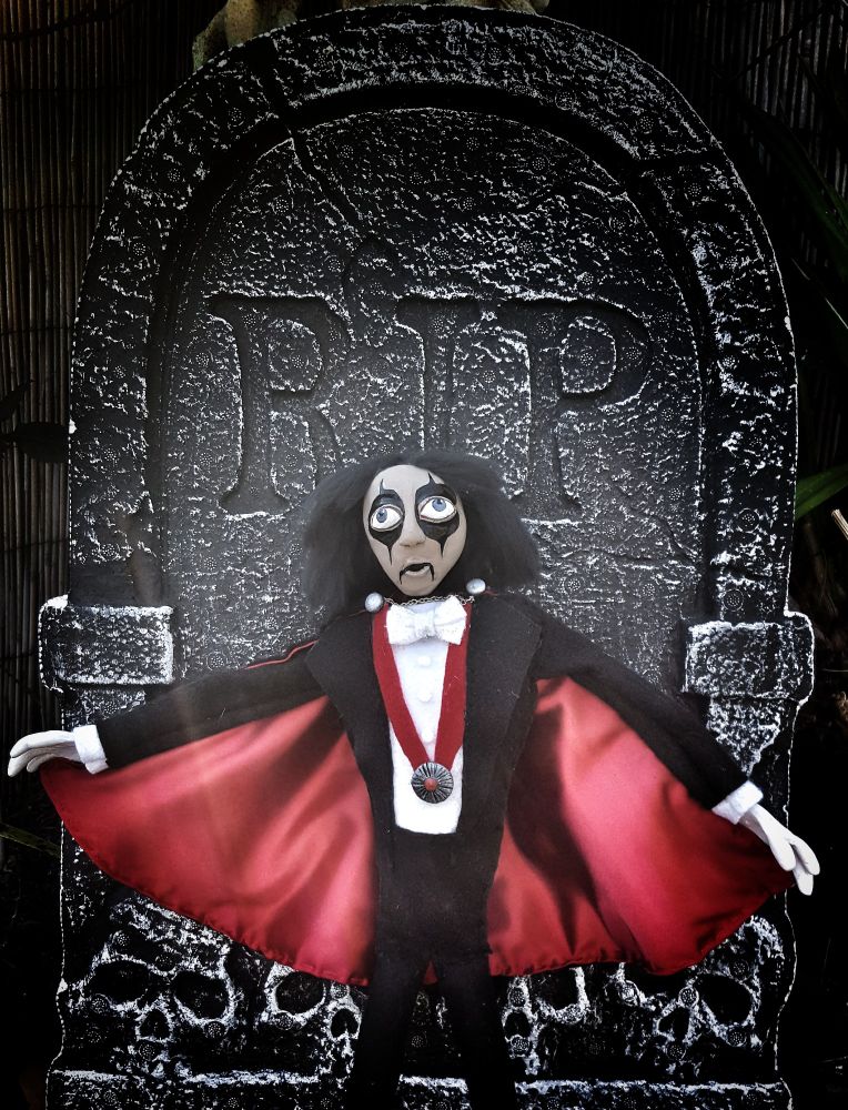 Doll of Alice Cooper standing in front of faux tombstone.