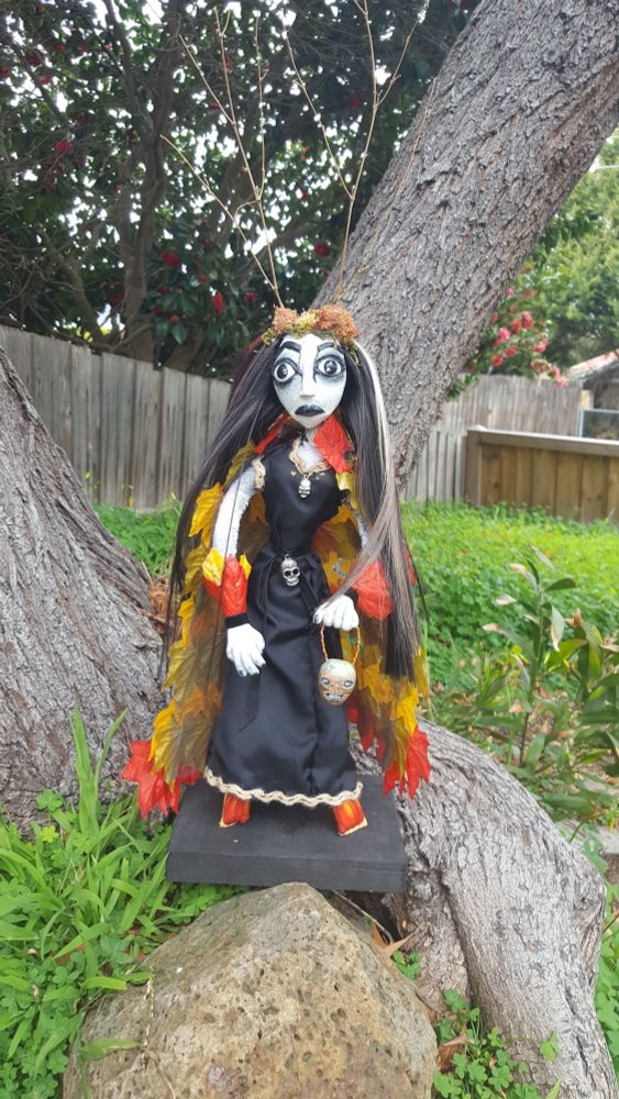 Photo of a doll standing on a rock, with a tree trunk with grass and a fence in the background.