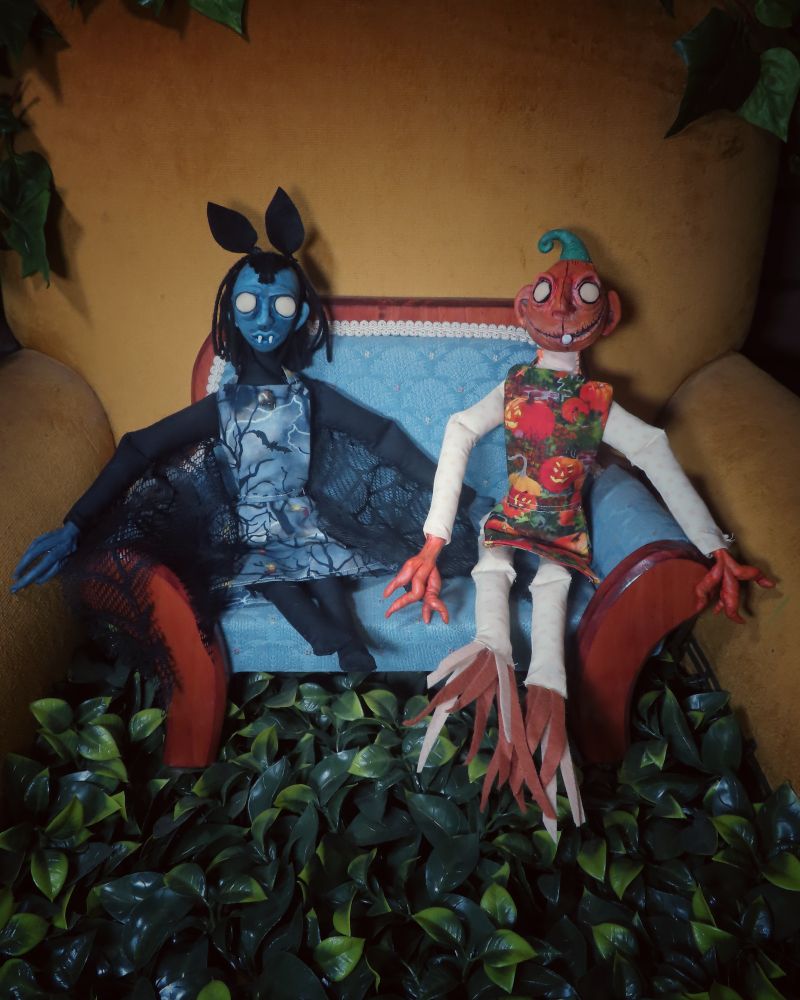 Yellow chair with faux greenery. On the chair is a brown and blue doll couch. On the couch are two dolls - on the left the doll is bat themed, on the right the doll is jack o lantern themed.