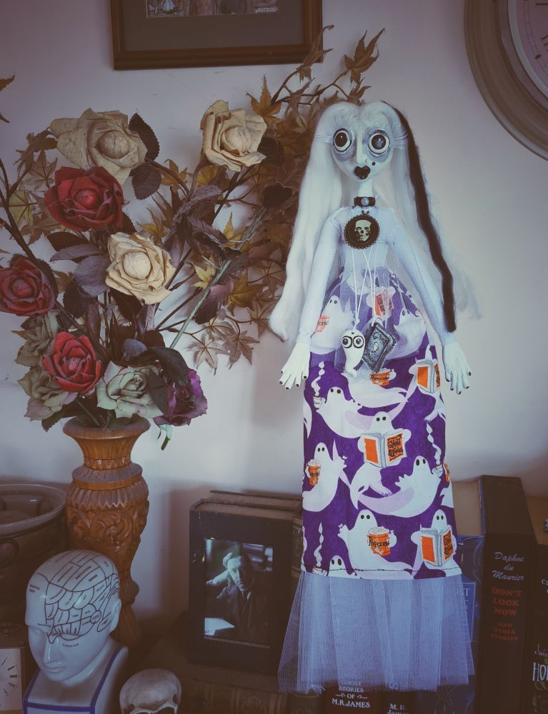 Doll on a shelf, surrounded by books, prints on the wall, a sculpture and a wood vase with faux flowers. 
