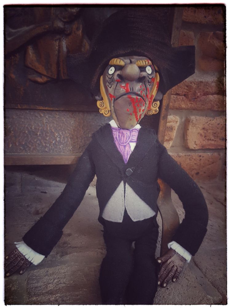 Doll stands in front of brick wall and metal fireplace screen.