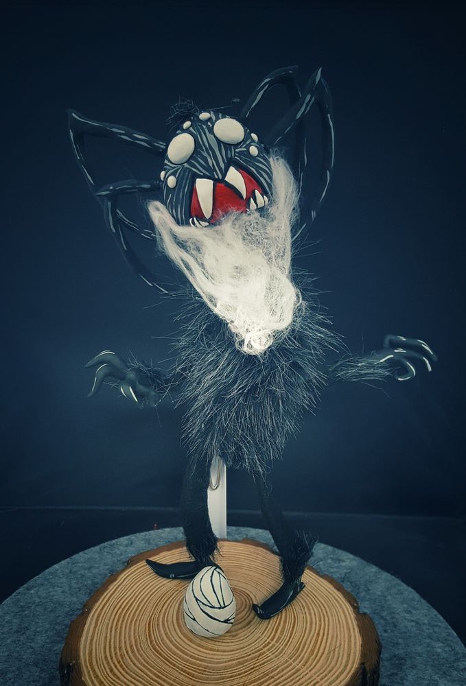  A fuzzy furred doll with a spider head and webbed beard stands on a wood disk, which is on a round grey felt mat. At the dolls feet is a sculpted spider egg.