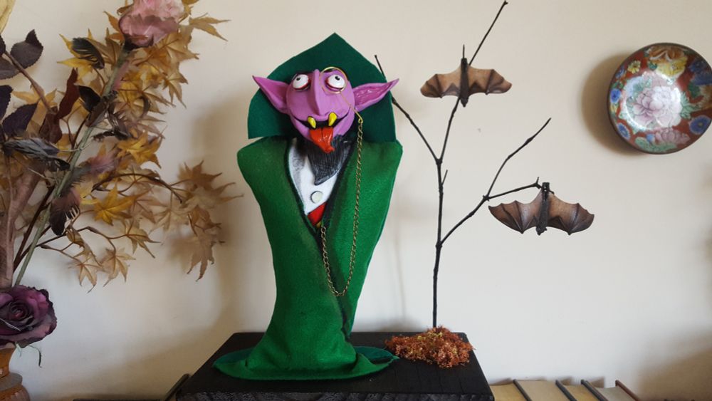 Sculpture of The Count with faux tree, bats and greenry on a wood block. Beside him is more faux greenery and  decorative plate on a wall.