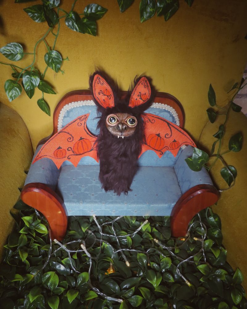 Photo of a yellow chair with faux greenery. On chair is a doll couch. On the doll couch is a bat push with pumpkins painted on the wings.