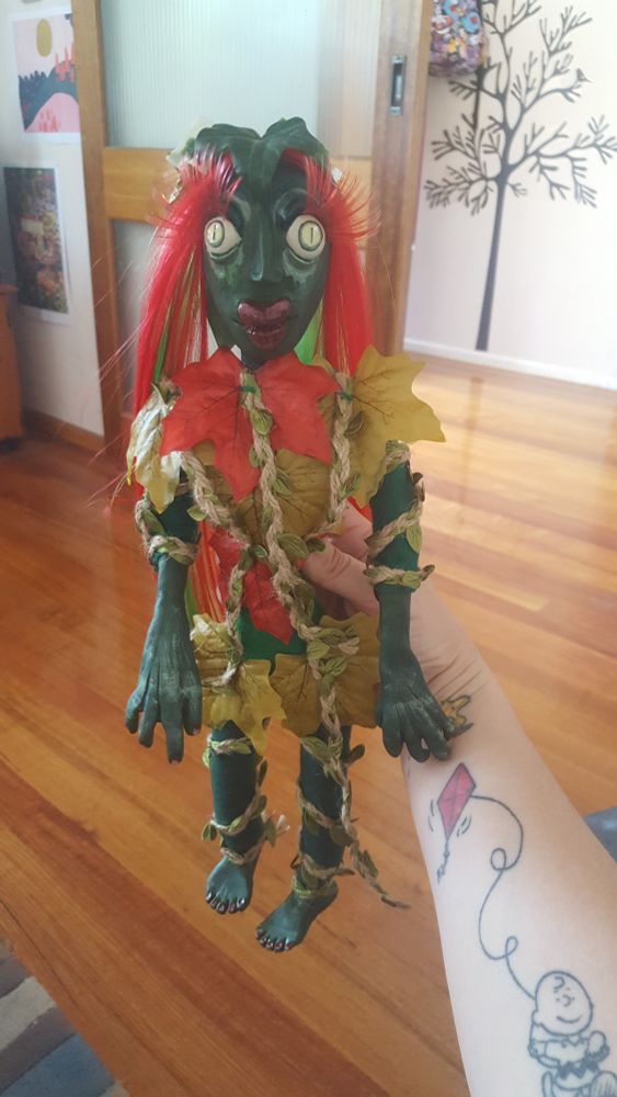 Strange doll of Poison Ivy beig held up, with wood floor, wall and sliding glass door in background.