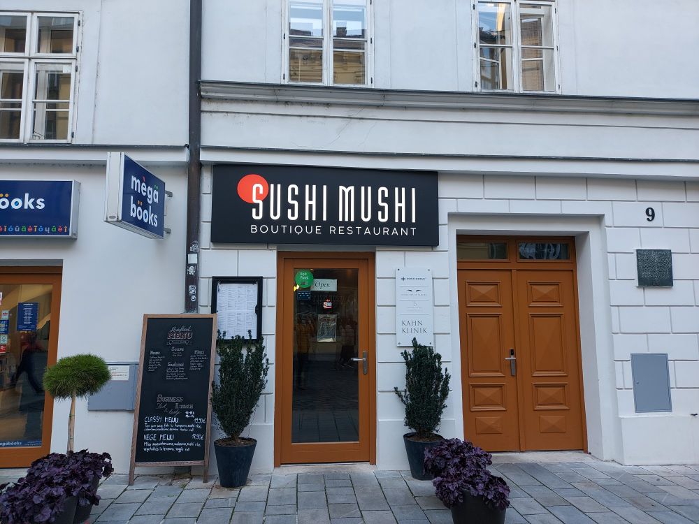 Weiße Hausfassade mit schwarzem Schild über einer Glaseingangstür mit braunem Rahmen. Auf dem Schild steht: "SUSHI MUSHI BOUTIQUE RESTAURANT".