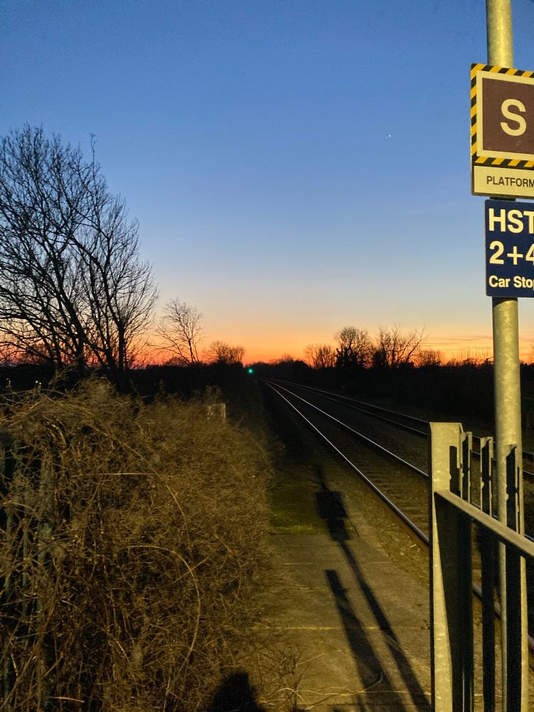 View of a railway line at late dusk at a rural station.  The railway track is light by the electric light from the platform.  