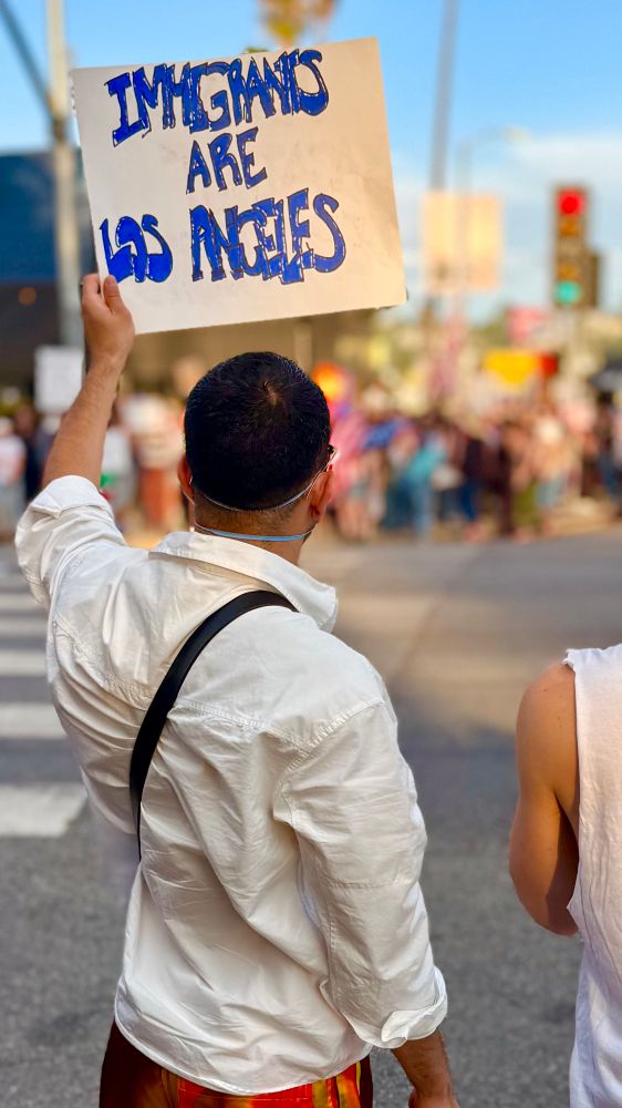 A masked protester holds a sign that reads Immigrants ARE Los Angeles; a crowd is visible across the street 