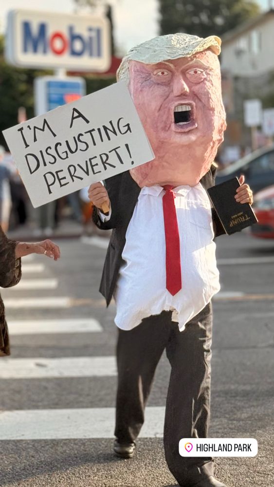 A protester dressed sloppily with a giant paper mache head of Trump, carrying a sign that says I’m a disgusting pervert