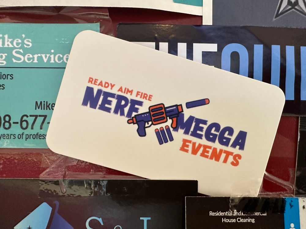 A business card on the bulletin board of a local deli, advertising “NERF MEGGA Events”. The words “NERF MEGGA” are divided by a drawing of a nerf gun, at an angle, which is covering part of the M. At a glance, it appears to say “NERF (N-Word)”