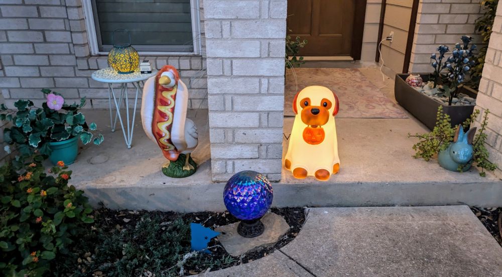 a lawn goose dressed in a wonderful hotdog costume next to a lit up plastic dog dressed up for Halloween 
