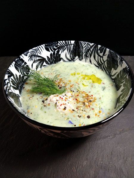 Auf einer Schieferplatte vor dunklem Hintergrund steht eine weißgrundige Schale mit schwarzem Blättermuster. Darin zartgrüne Cremesuppe, auf der Oberfläche ein heller Klecks Rahm, grüne Dillspitzen, einige Tropfen goldgelbes Öl und grob geschrotete bunte Gewürze.
