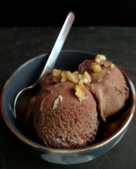 In einer kleinen dunkelgrauen Dessertschale vor dunklem Hintergrund sind zwei braune Kugeln Eiscreme, bestreut mit gehackten Walnüssen.