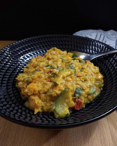 Auf Holzfläche vor dunklem Hintergrund ein schwarzer Teller mit einer Portion gelbem Linsencurry. Es sind grüne Zucchinistücke und kleine rote Chili- und Paprikawürfel darin zu sehen.