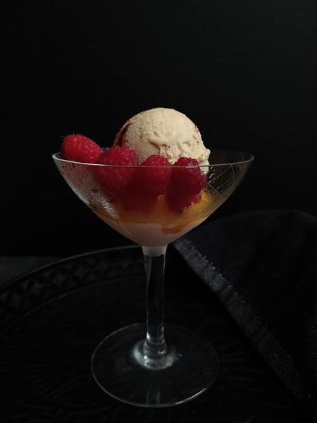 Dunkler Hinter- und Untergrund. Ein 50er-Jahre Cocktailglas, von der Kälte leicht beschlagen. Am Boden des Glases eine orangene Schicht Nektarinenröster (Kompott ohne Flüssigkeitszugabe). Darauf eine cremefarbene Kugel Eiscreme, am Rand ist eine kurze Linie vom darübergeträufelten Balsamico zu sehen. Daneben sind einige rote Himbeeren arrangiert.