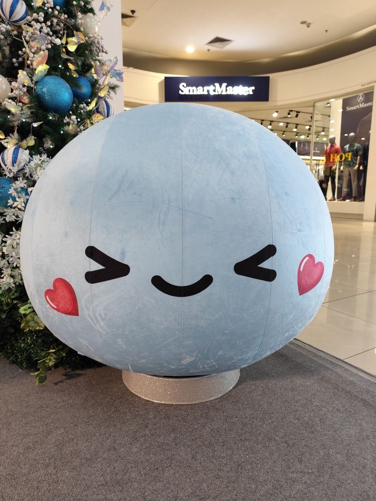 Photo of a balloon of the light blue mochi mascot of AEON Mall in a Santa hat with a smiling squinting face and heart-shaped blushes, positioned next to a Christmas tree.