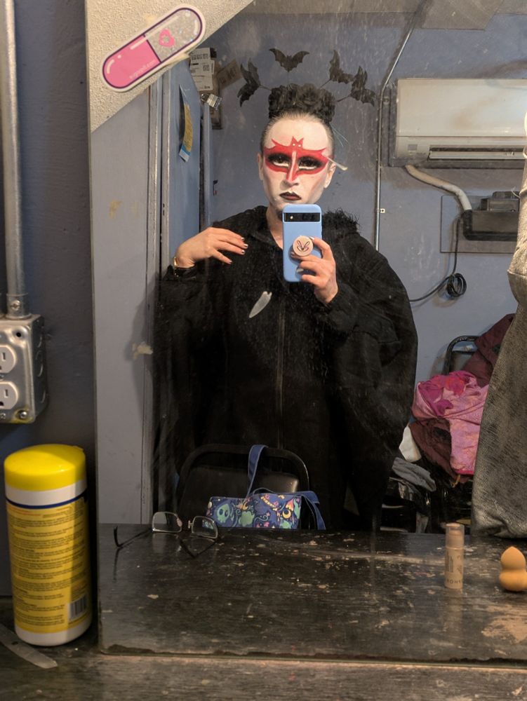 Jaggs Ramone, a drag king, wearing a black outfit and a headband that has bats on it. his makeup is corpse paint style, with white foundation and red paint styled to look like a bat with spread wings