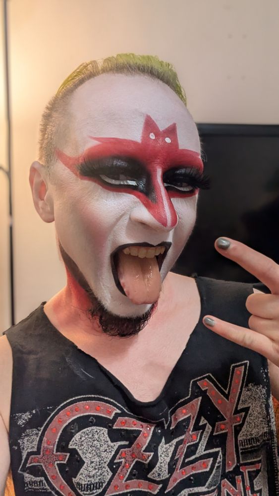 a drag king in a rhinestoned Ozzy shirt and corpse paint inspired makeup styled to look like the silhouette of a bat. he has his tongue sticking out and is displaying the devil horns with one hand
