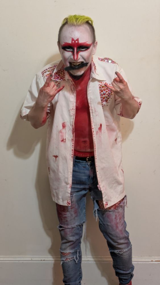 same king, but we can see more of his outfit, including torn and stained jeans. both hands are displaying the devil horns, and there's a rubber bat in his mouth 
