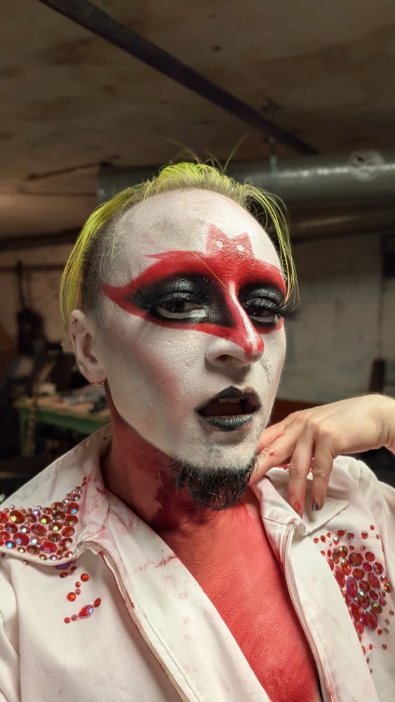 same king, but in a white lab coat thats been rhinestoned to look like blood has soaked into it. his makeup has worn away slightly at the mouth and his chest is covered in red cosmetic paint
