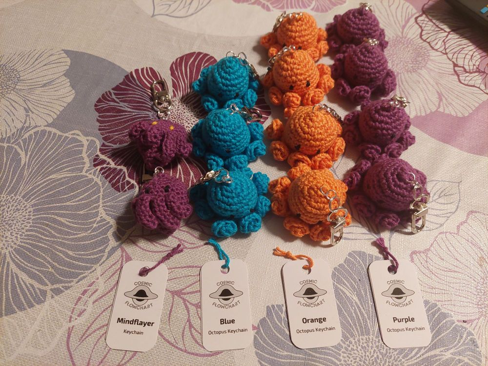Four rows of crochet keychains/bag charms. In the leftmost row, there are two purple keychains shaped like the head of a Mindflayer from Dungeon and Dragons, with four tentacles. One has black eyes, the other yellow. The remaining keychains are octopus shaped and have black eyes.  From left to right, their colors are: blue, orange and purple. Below each row is a card stock tag. Each shows a grayscale version of my logo, an accreting black hole with the word "cosmic" above and the word "flowchart" below it. The remaining text on the leftmost tag reads "Mindflayer" in bold and "Keychain" in a smaller, regular font. Similarly the remaining three tags state the octopus color in bold, i.e., blue, orange, and purple, respectively. The smaller font on all three tags reads "Octopus keychain". 