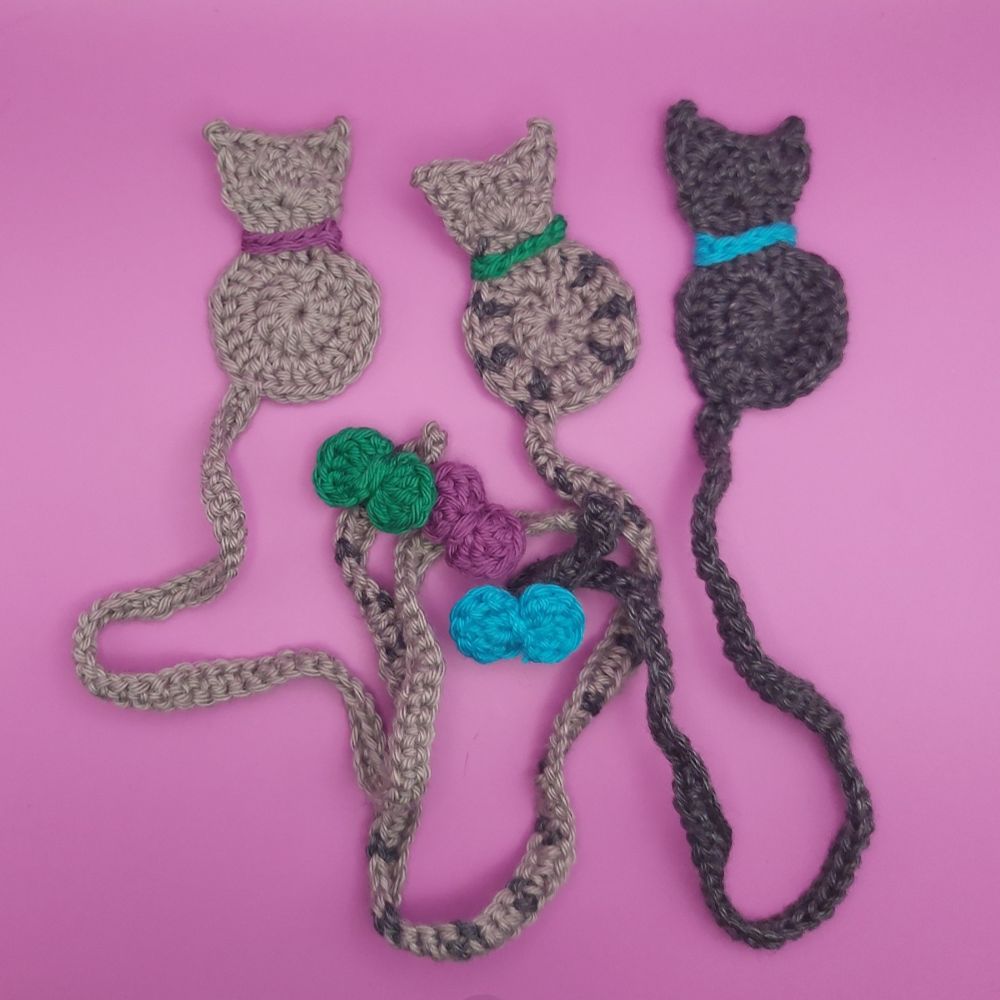 Three crocheted bookmarks shaped like cats. Each has a collar and a bow on the tail as accessories. The left bookmark is light gray with purple accessories, the right one is dark gray with light blue accessories, and the one in the middle light gray with dark gray stripes on the side and tail with green accessories.