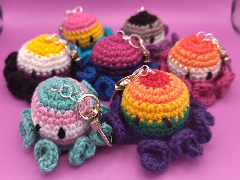 Crochet keychains shaped like octopuses, with colorful stripes matching various LGBTQIA+ pride flags, rainbow, bisexual, transgender, pansexual, non-binary, asexual, and lesbian.