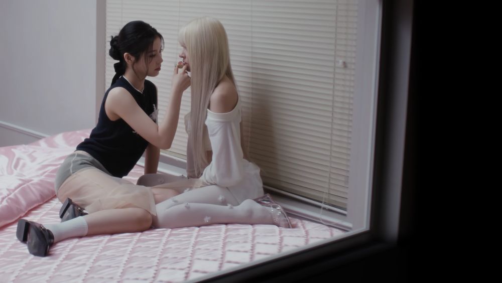 screencap showing nmixx's kyujin putting something against lily's lips. they are sitting in a room with blinds on one side and a pink duvet on the floor