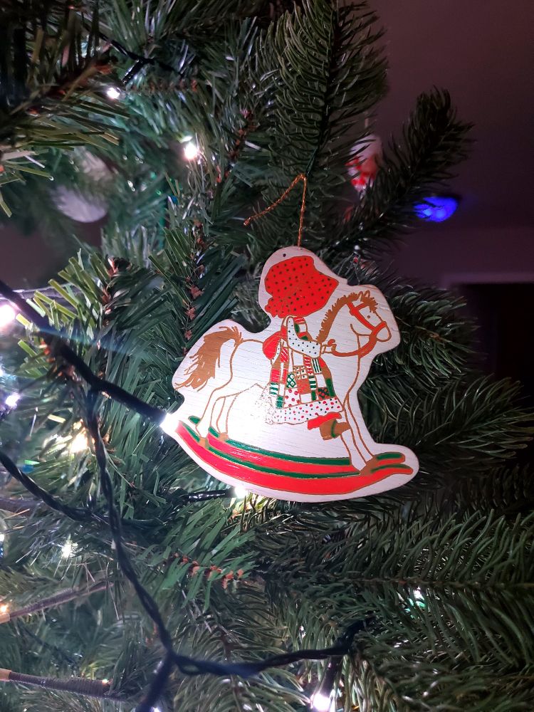A wooden ornament with a painting of Holly Hobbie (a child in profile wearing a red, green, and white patchwork dress, boots, and a large red Polka dot bonnet that blocks her face) riding a white rocking horse.