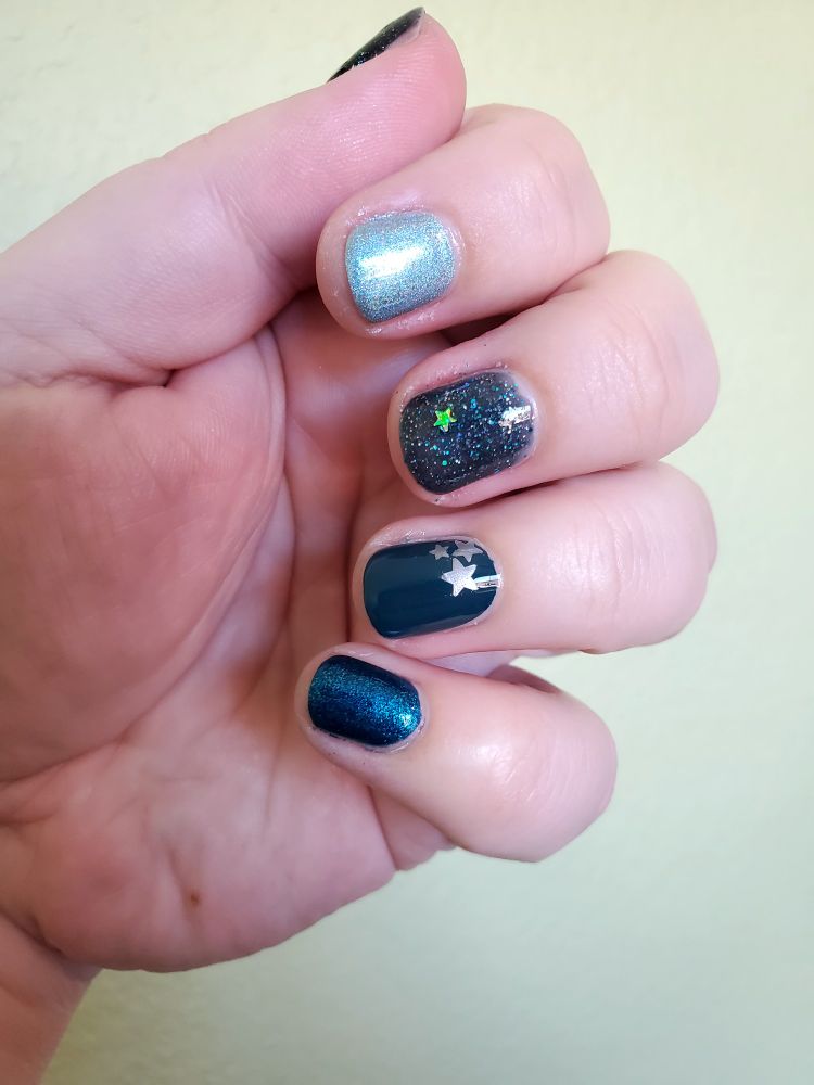 A white lady's hand with a manicure in shades of a bright holographic metallic aquamarine, teal with layers of holographic sparkles and a silver star, a deep teal creme with 3 silver stars in one corner, and a shimmery blue teal.