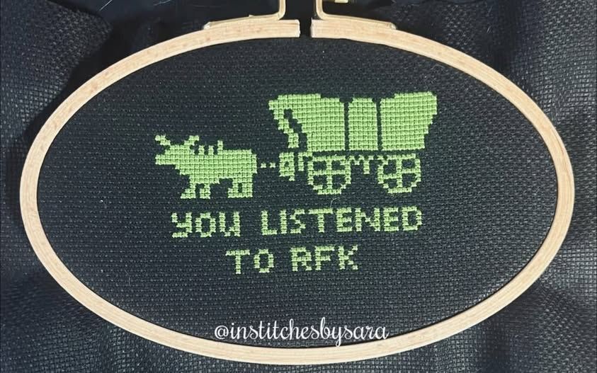 A cross stitch design in light green on a black background, framed with a tan oval embroidery hoop. The stitching shows a very recognizable ox-drawn covered wagon from the "you have died" messages in the computer game Oregon Trail, with the text "You listened to RFK". The watermark on the image is @institchesbysara