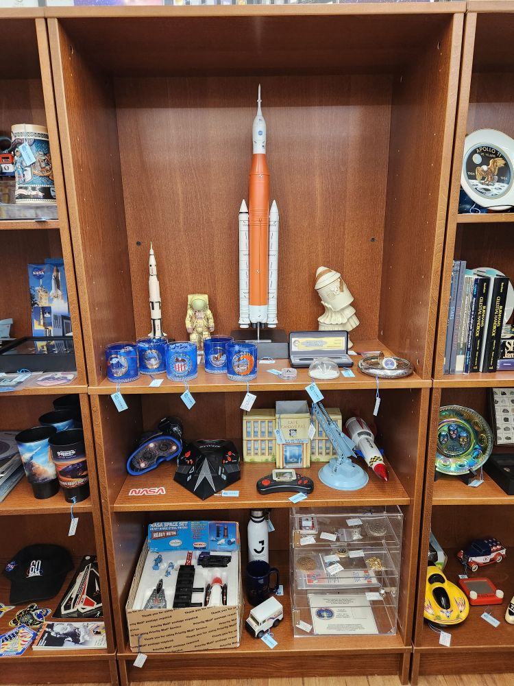 Photo of back wall (center bookshelf)  of antique mall booth that highlights various space collectibles, including a large scale SLS rocket and other various vintage space toys.