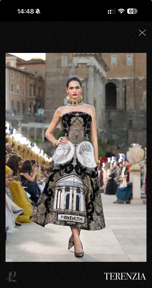 A model walks down a runway wearing an elaborate black strapless gown with ornate gold embroidery and decorative motifs. The dress features classical architectural imagery including the Pantheon, along with coin portraits and intricate baroque-style patterns. She’s wearing a statement gold collar necklace and has her dark hair pulled back. The runway show is taking place in an Italian piazza with classical stone architecture visible in the background. Seated spectators line both sides of the runway. The image has “TERENZIA” credited at the bottom.​​​​​​​​​​​​​​​​