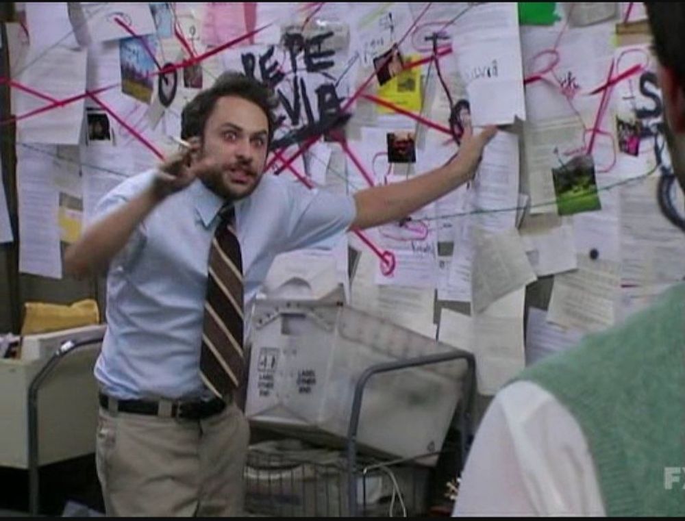 Homem aparentemente nervoso apontando pra um mapa cheio de fotos e riscos como se estivesse explicando uma trama ,ele está de camisa azul ,gravata vinho com listras brancas e calça caqui e segura um cigarro na mão.