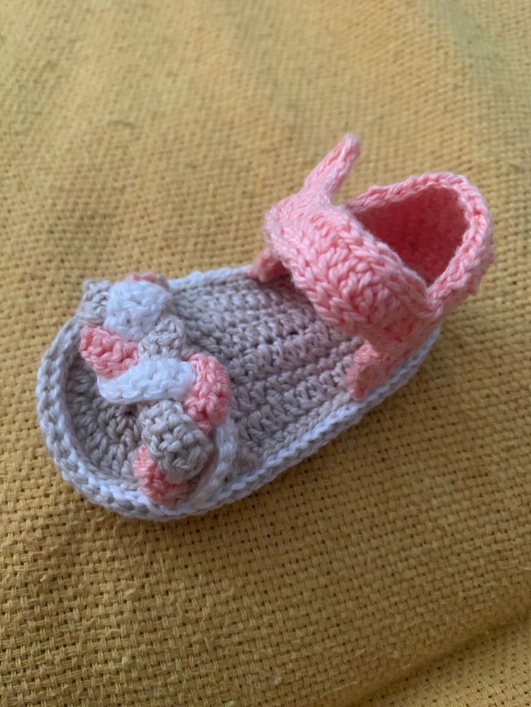 Sapatinho de crochê na cor bege, rosa e branco, na frente tem um tira de trança com três cores, e atrás é uma parte de abotoar está sobre um coxa amarela