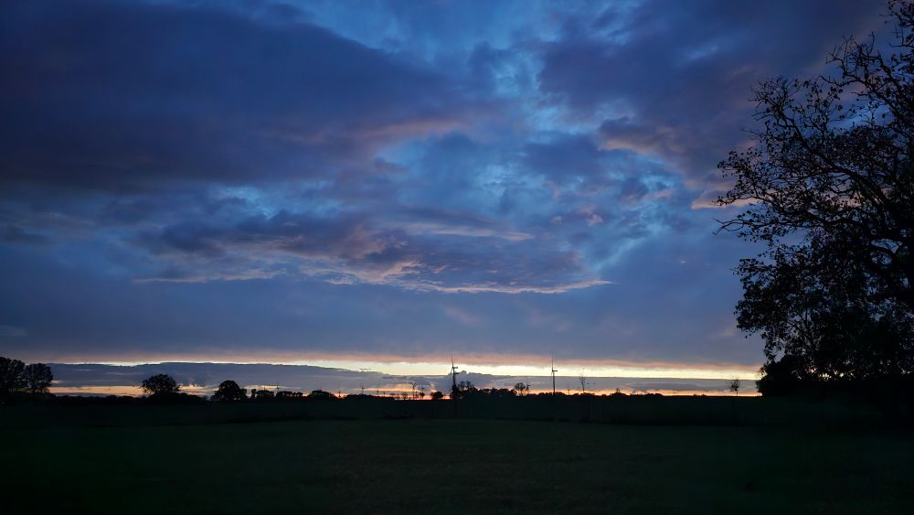 Die Sonne ist untergegangen an der Ostseeküste. Auf dem Foto sieht man ins Landesinnere auf dunkle Wolken und darunter ein helles Lichtband, in dem Windräder langsam vor sich hin drehen. 
